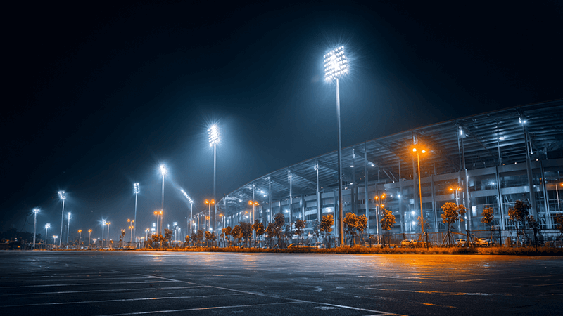 A modern stadium lit with bright, clear LED floodlights next to an old parking lot with yellow, dim traditional lights.